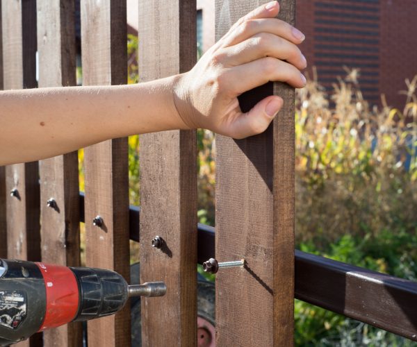 Close up of hand screewing wood plank to metal construction. Building a wooden fence with a drill and screw.